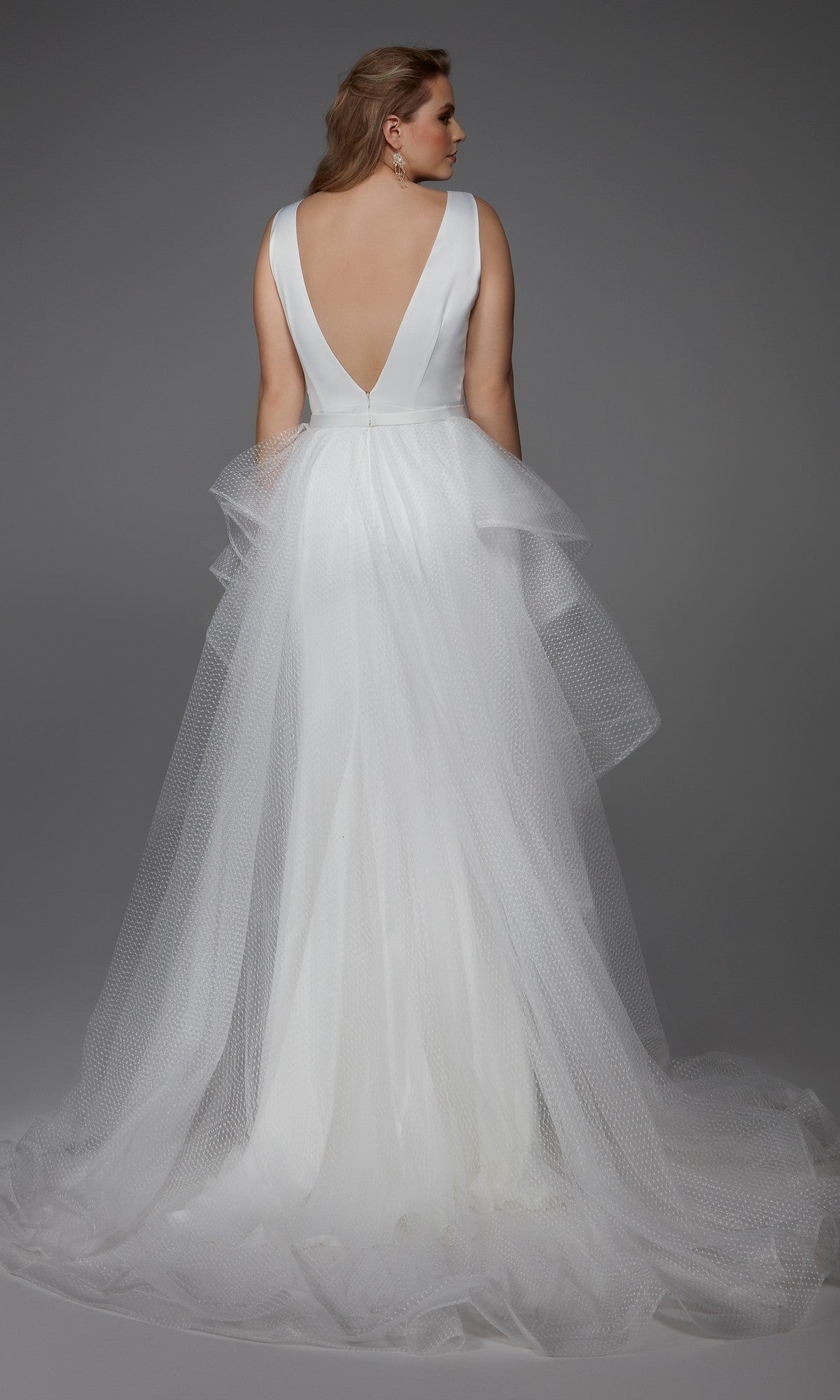 A woman wearing the Alyce 7019 long white dress—a sleeveless bridal gown with a deep V-back and layered tulle skirt—stands against a gray background, facing away from the camera.